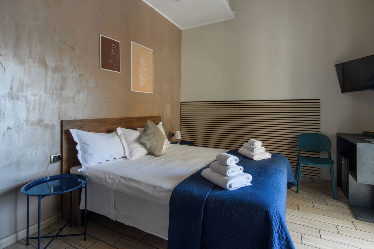A hotel room with a bed, a blue side table, a chair, a desk, and a television.