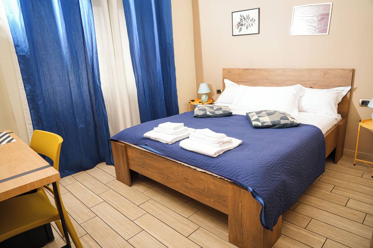 A neatly made bed with a blue quilt, white pillows, and folded towels. There is a wooden nightstand with a lamp, a yellow chair, and a small table with a lamp. The room has wooden flooring and blue curtains.