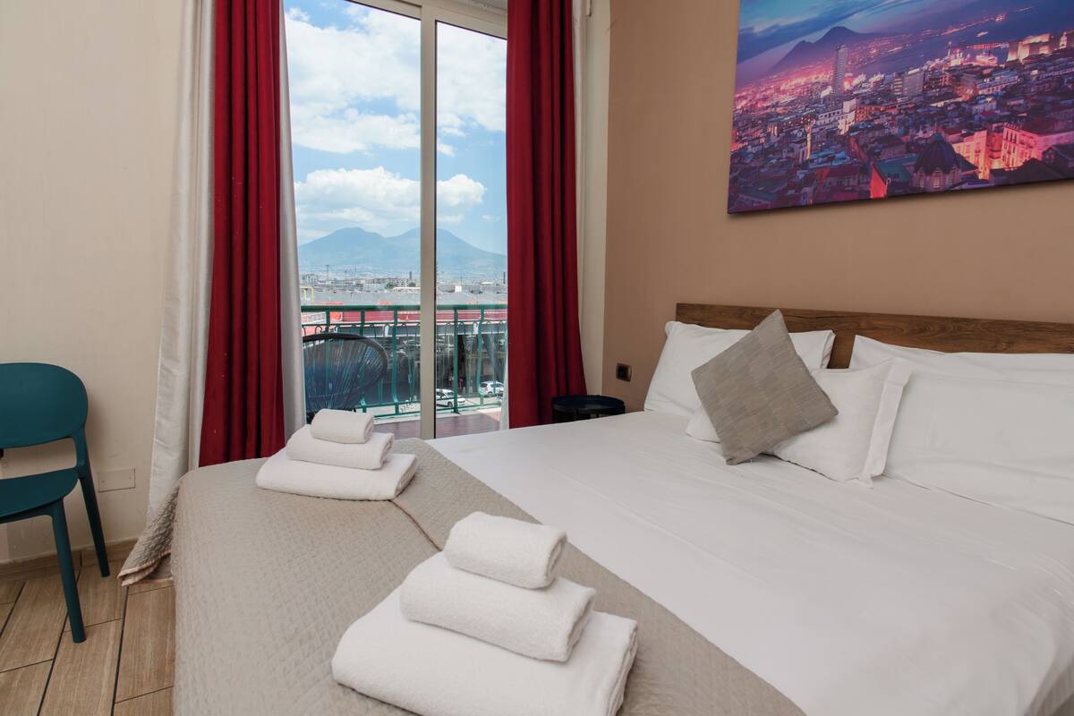 A hotel room with a large bed, a view of a cityscape, and a balcony with red curtains.