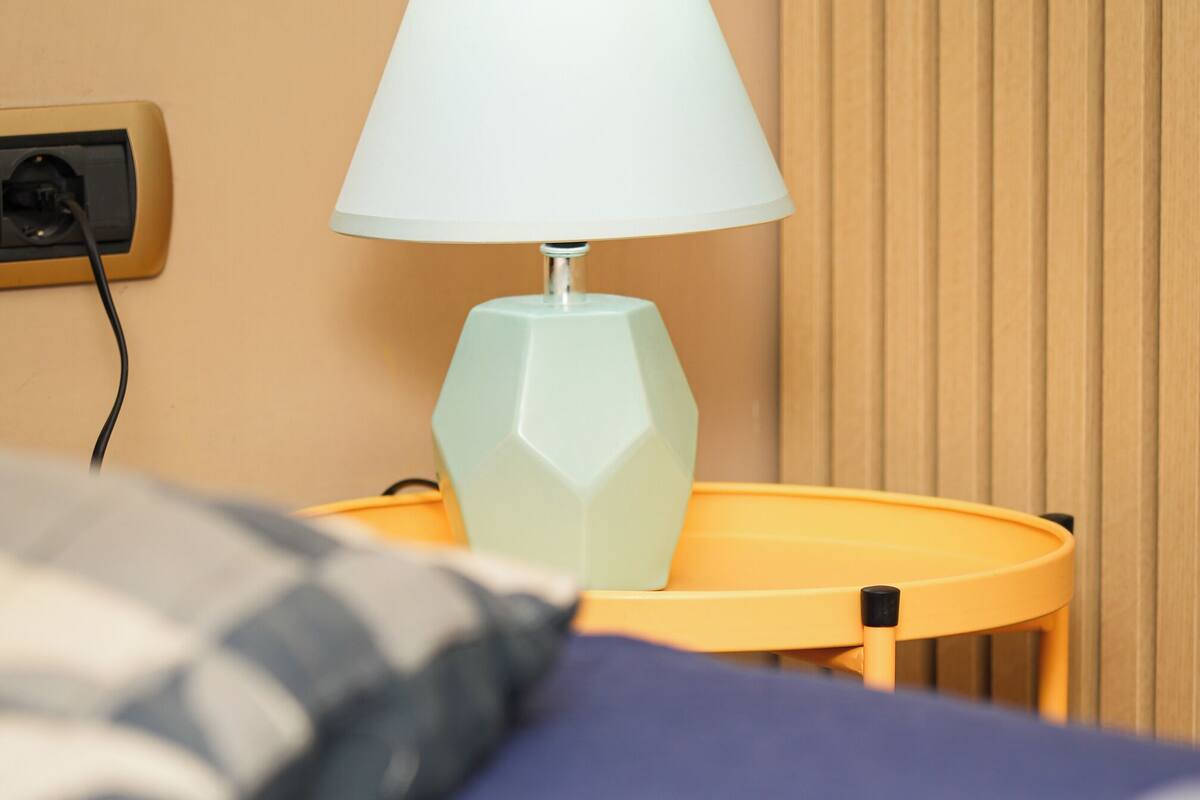 A bed with a blue bedspread, a light blue table lamp, and a pillow on a yellow side table.