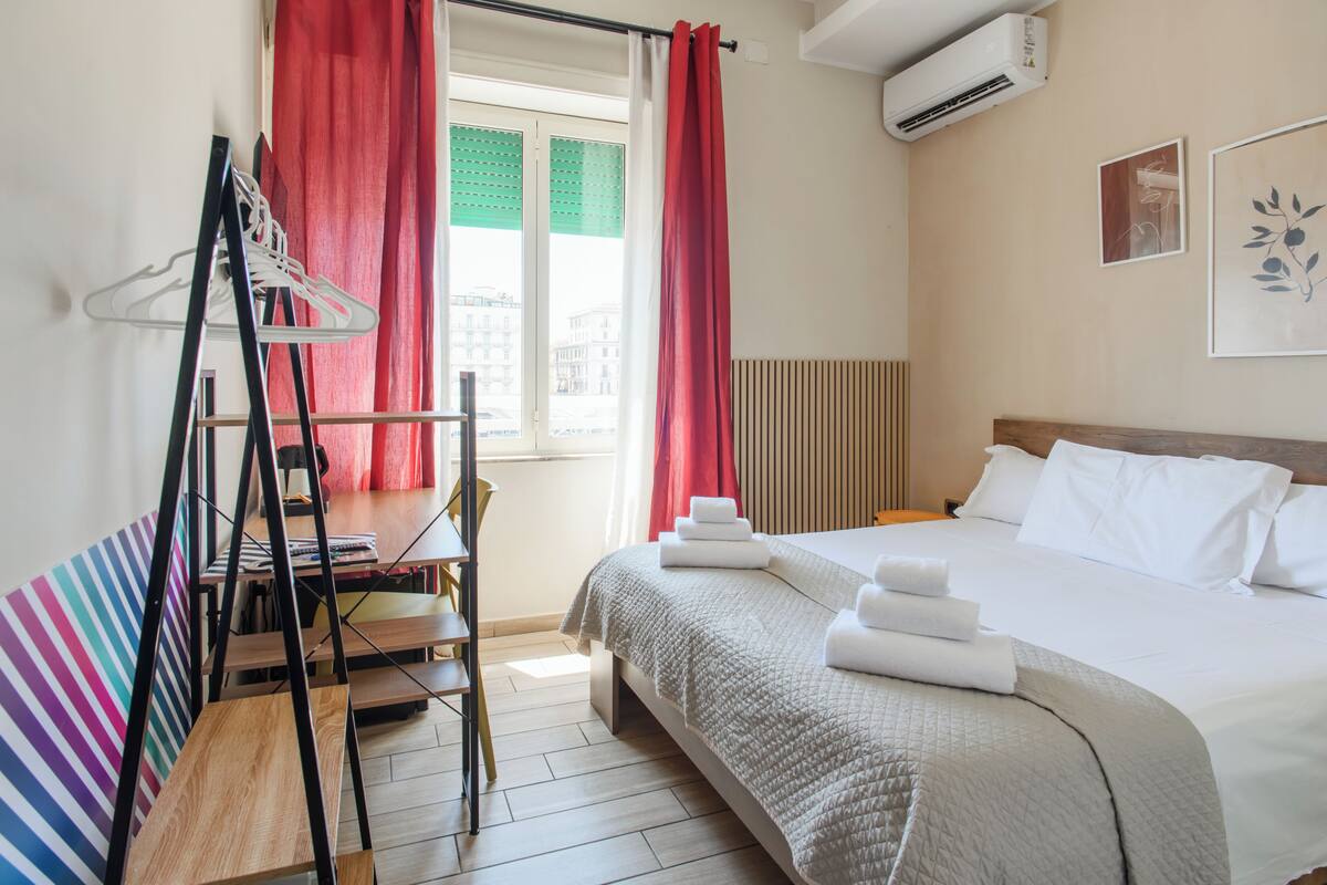 A hotel room with a bed, a desk with a chair, a window with curtains, and an air conditioning unit.