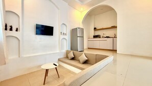 Un salon moderne avec un canapé, une petite table et un téléviseur. Au fond, on aperçoit une cuisine équipée de placards et d’un four à micro-ondes.