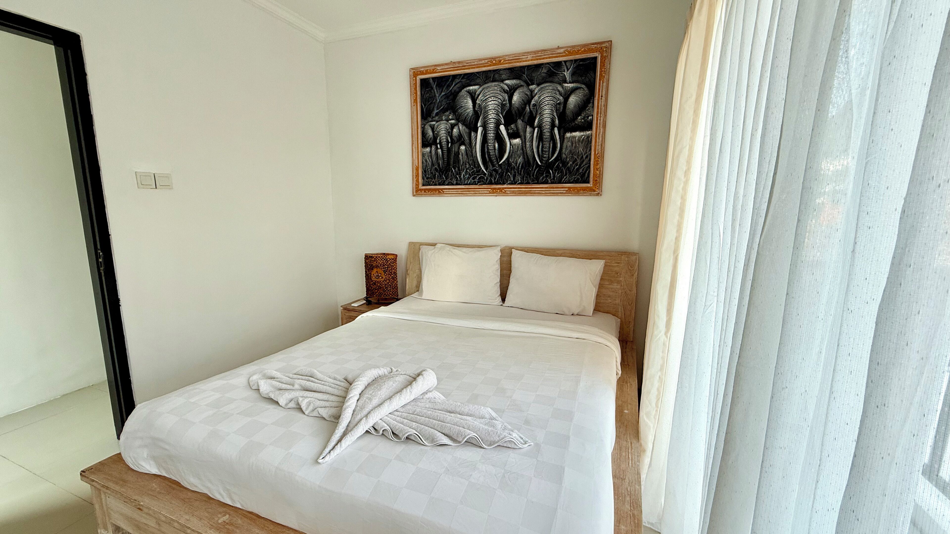 A neatly made bed with a checkered bedspread and a folded towel, a wooden headboard, a nightstand with a lamp, and a framed artwork of elephants on the wall.