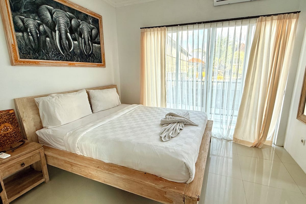 A neatly made bed with a checkered bedspread and a folded towel, a wooden nightstand with a lamp, and a painting of elephants on the wall.