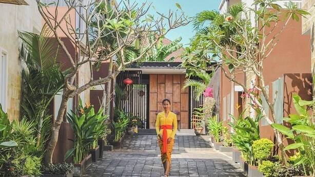 Une personne marchant sur un chemin bordé de plantes en pot et d’arbres, qui mène à l’entrée d’un bâtiment.
