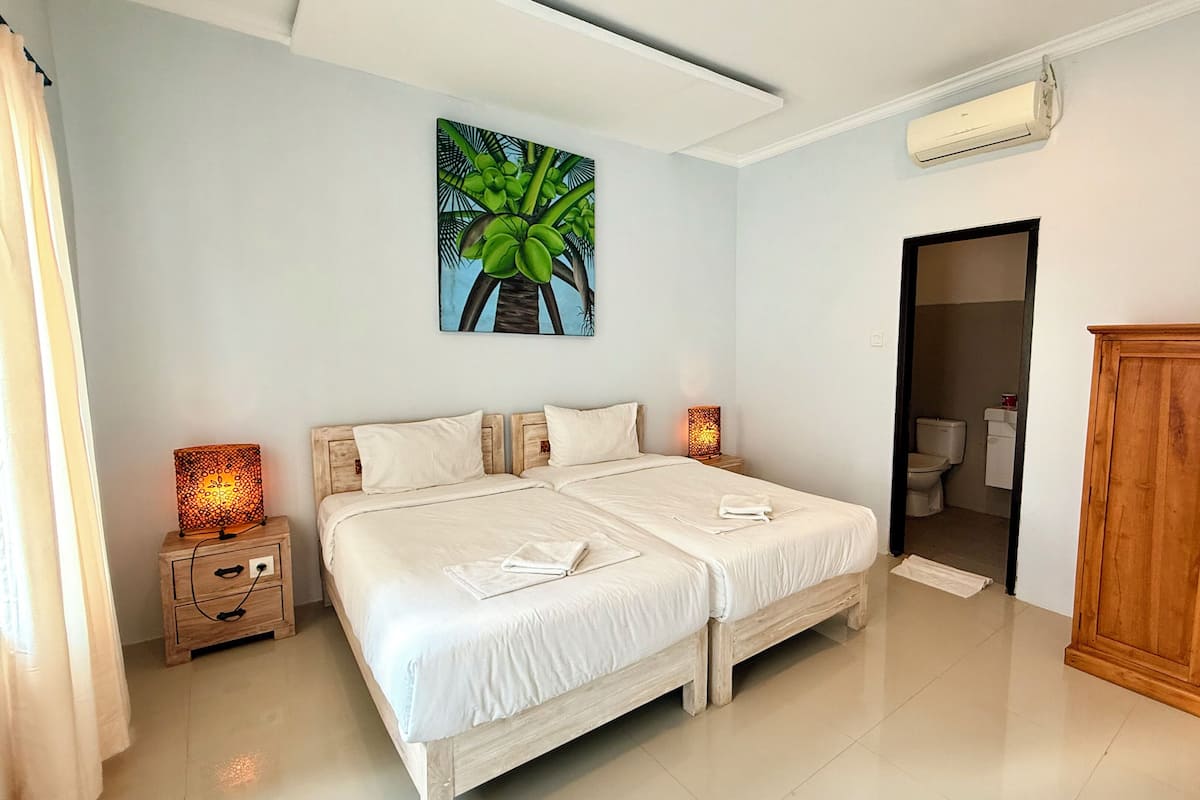 A bedroom with two beds, a painting of a palm tree, a wooden wardrobe, and a bathroom visible through an open door.