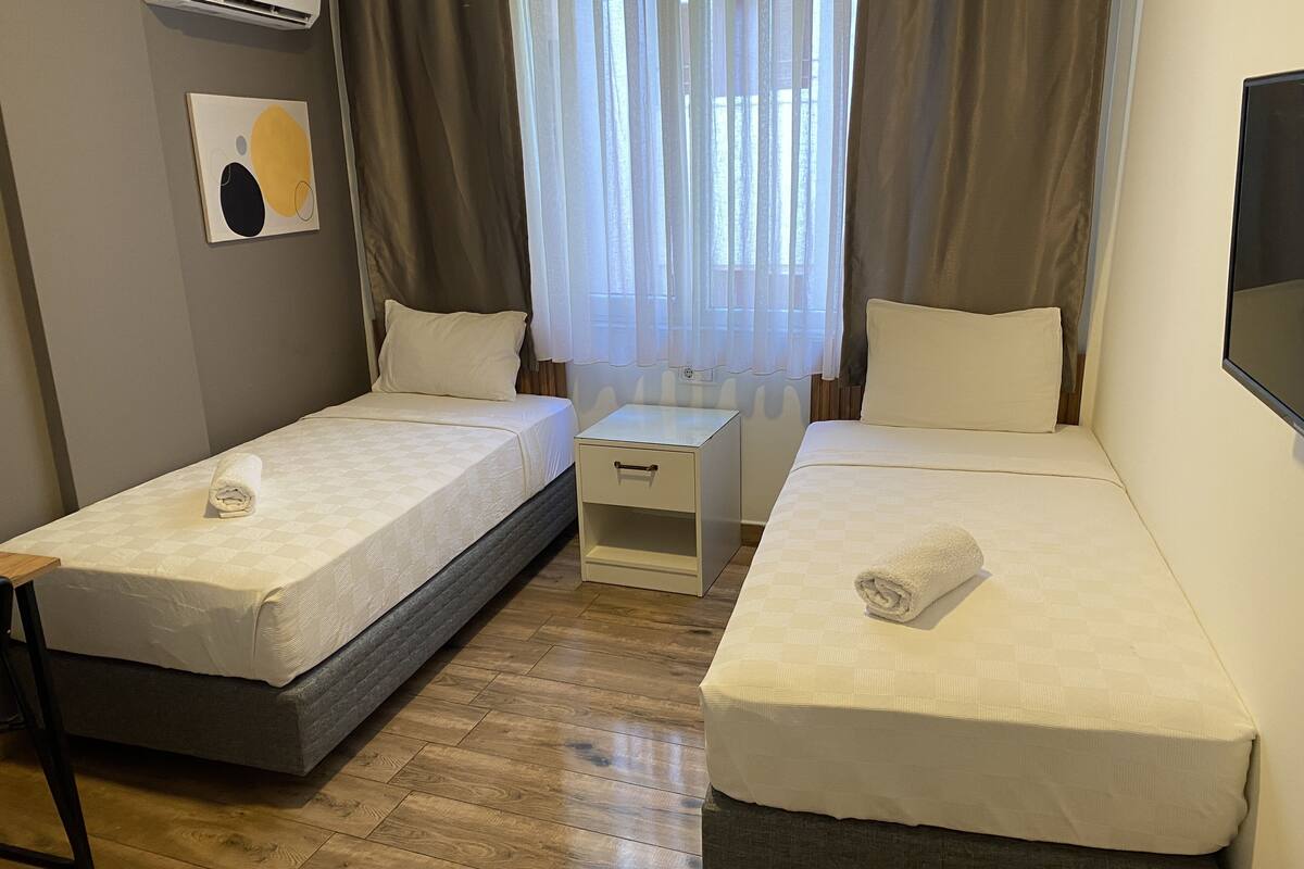 A twin room with two single beds, a small bedside table, a wall-mounted TV, and a window with curtains.