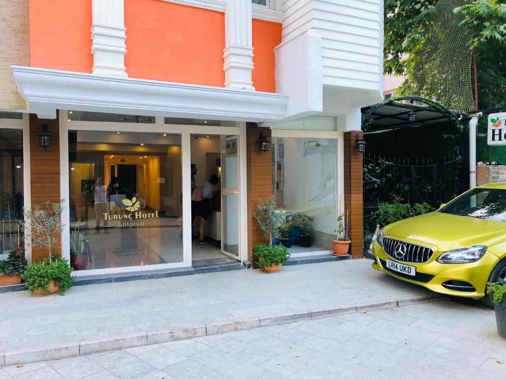 Entrada de un hotel con fachada de vidrio, un coche amarillo estacionado adelante y plantas en macetas afuera.