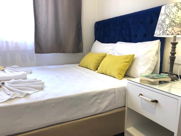 Una cama bien hecha con almohadas blancas y amarillas, una mesita de noche con una lámpara y una ventana con cortinas.