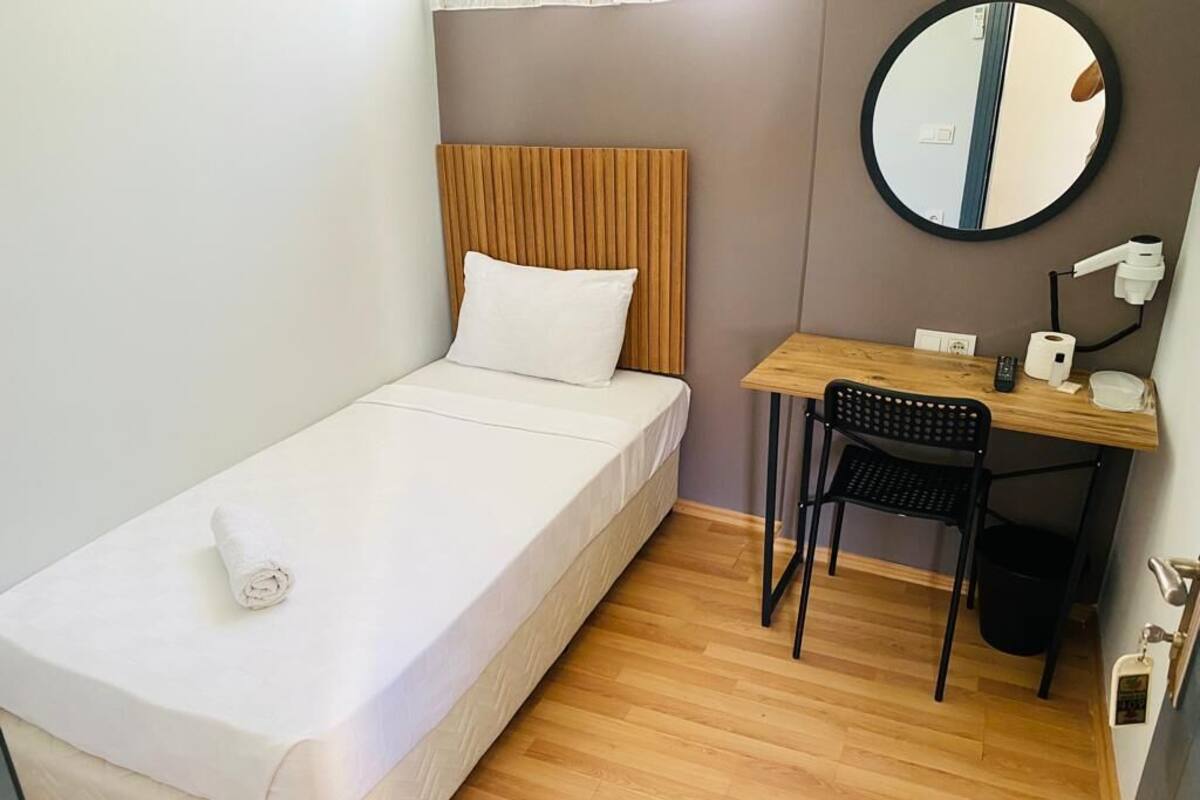A small, neatly arranged room with a single bed, a wooden desk, a chair, a round mirror, and a small table with a lamp and a remote control.