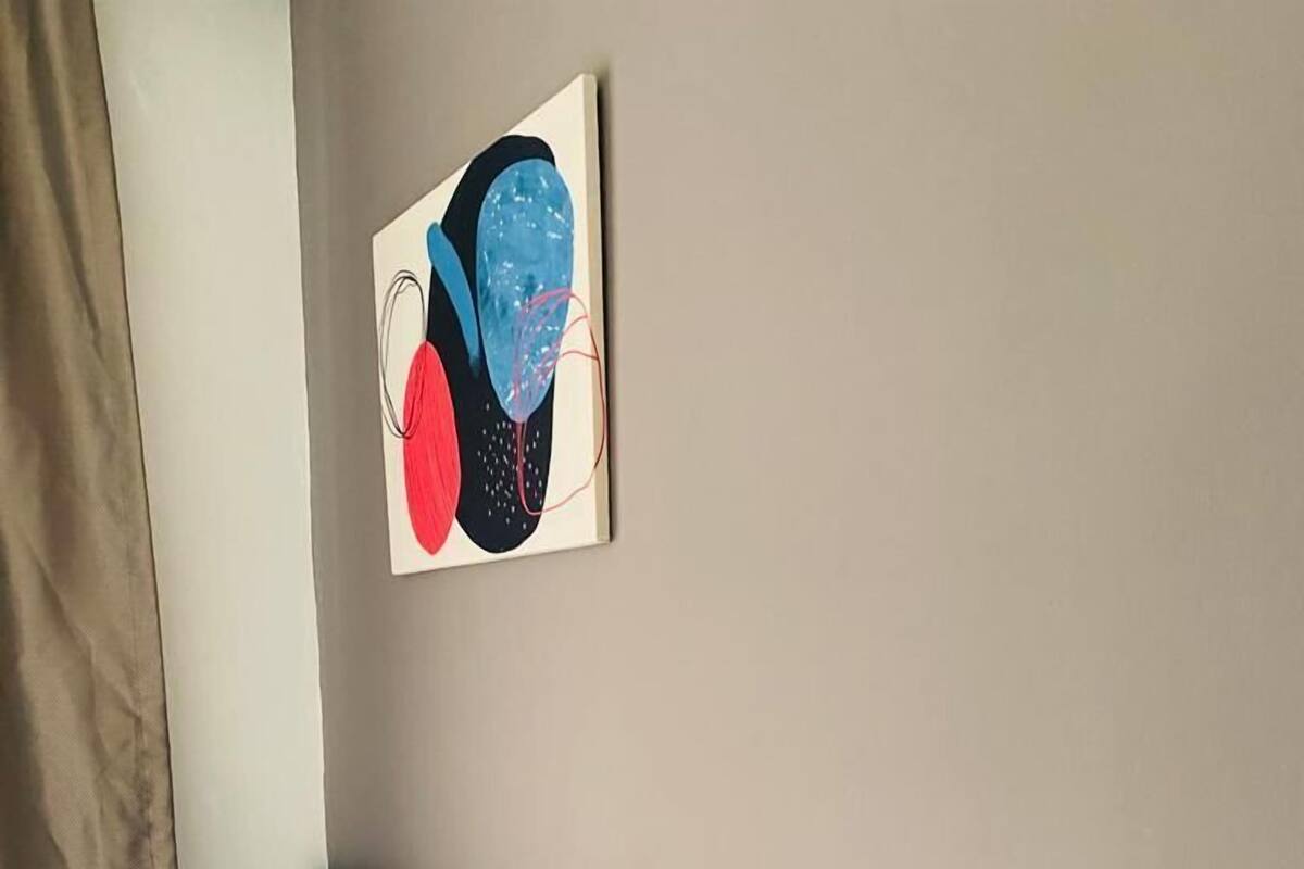 A hotel room with a blue tufted headboard, white bedding, a wall-mounted light fixture, and a piece of abstract art.