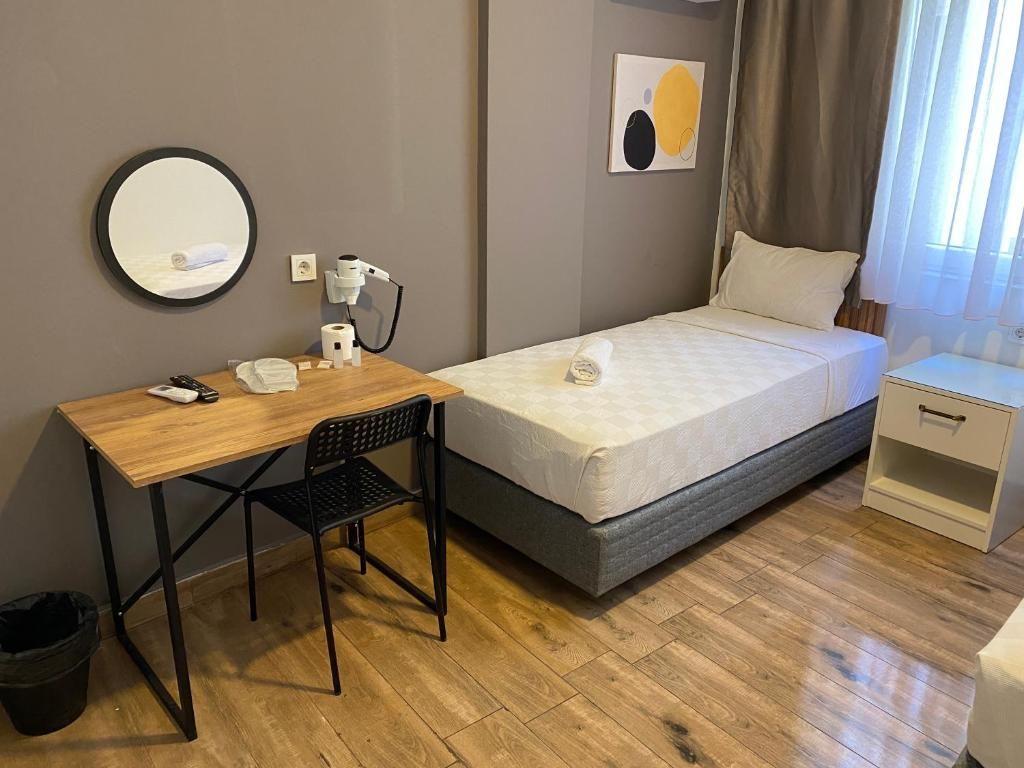 A bedroom with a bed, a desk with a chair, a bedside table, a mirror, and a wall-mounted artwork.