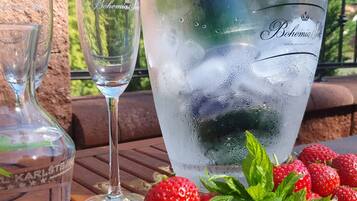 Una jarra de vidrio, una copa de vino, y un vaso grande con hielo y una pajita, sobre una mesa al aire libre con un banco de madera y un plato de fresas.