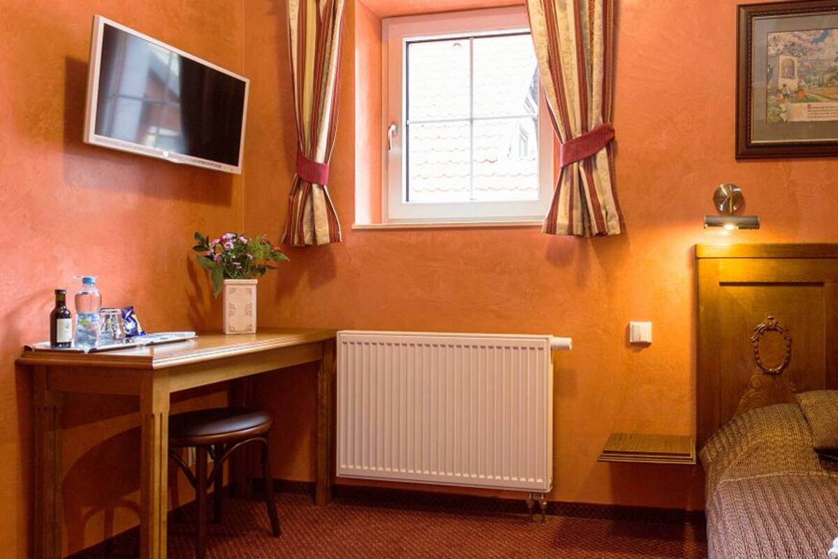 A bedroom with a bed, a desk, a radiator, a TV, and a window with curtains.
