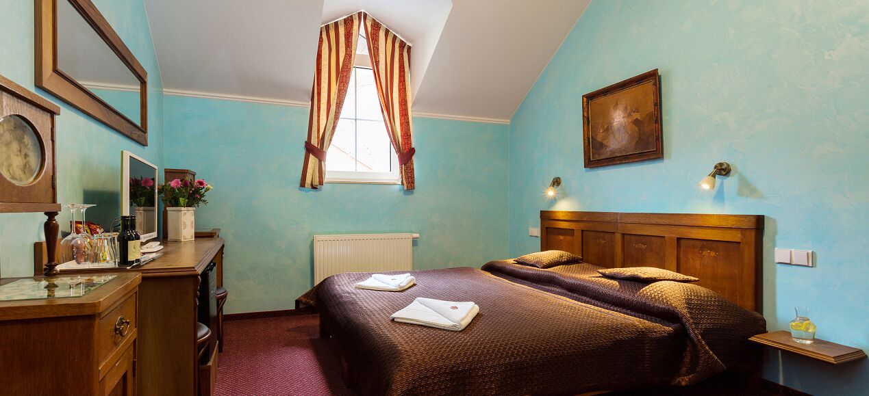 A bedroom with a bed, a desk, a chair, a radiator, a window with curtains, and a wall-mounted lamp.