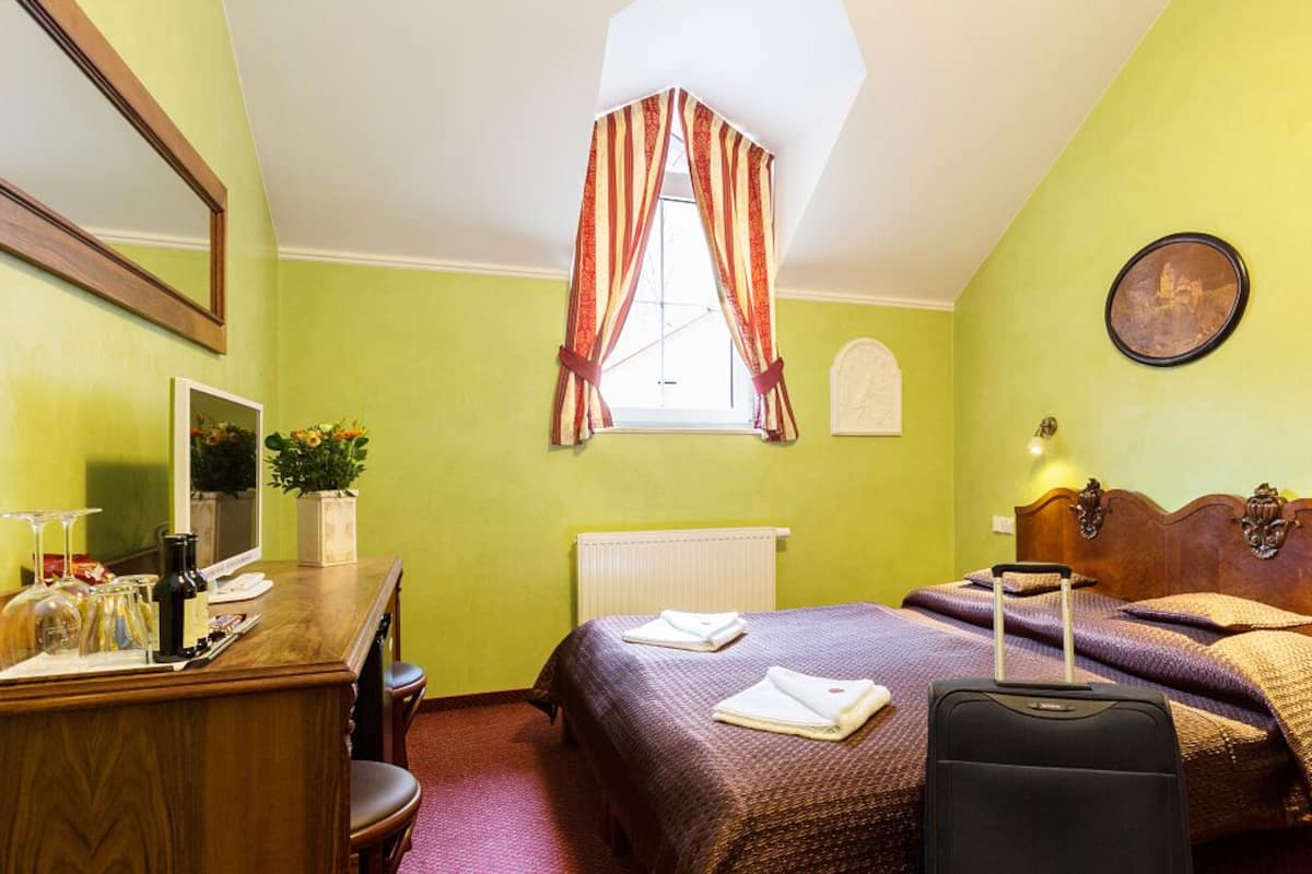 A hotel room with two beds, a desk with a chair, a radiator, a window with curtains, and a wall-mounted picture.