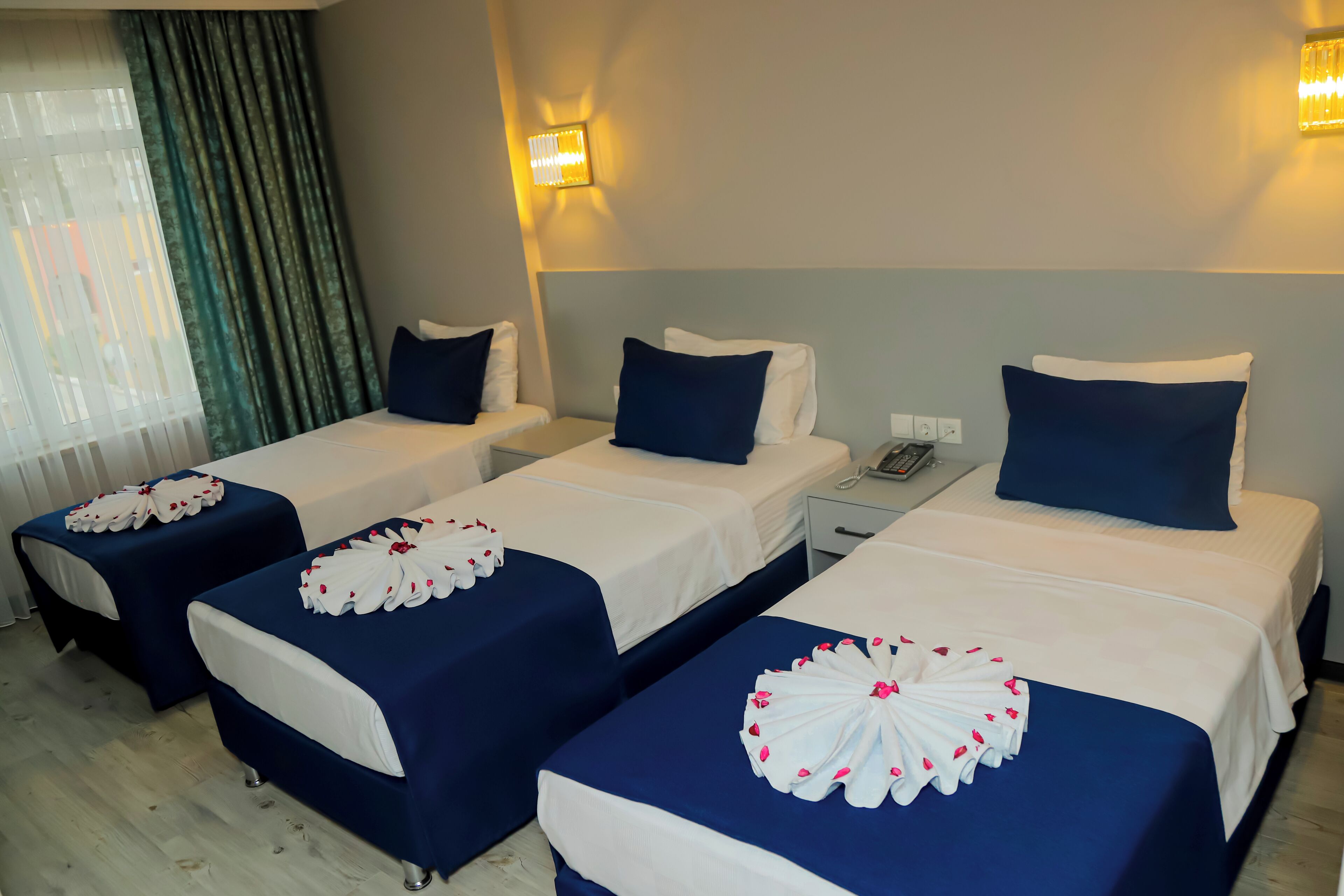 A hotel room with three single beds, each with a white and blue bedspread and a decorative white floral arrangement. There are two bedside tables with lamps, a nightstand with a phone, and a window with curtains.