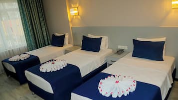 A hotel room with three single beds, each with a white and blue bedspread and a decorative white floral arrangement. There are two bedside tables with lamps, a nightstand with a phone, and a window with curtains.