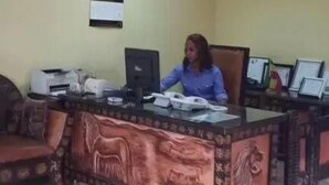 A woman sitting at a desk in a room with a tiled floor, a wall clock, and a framed picture. - The Lions Den Hotel (Addis Ababa)