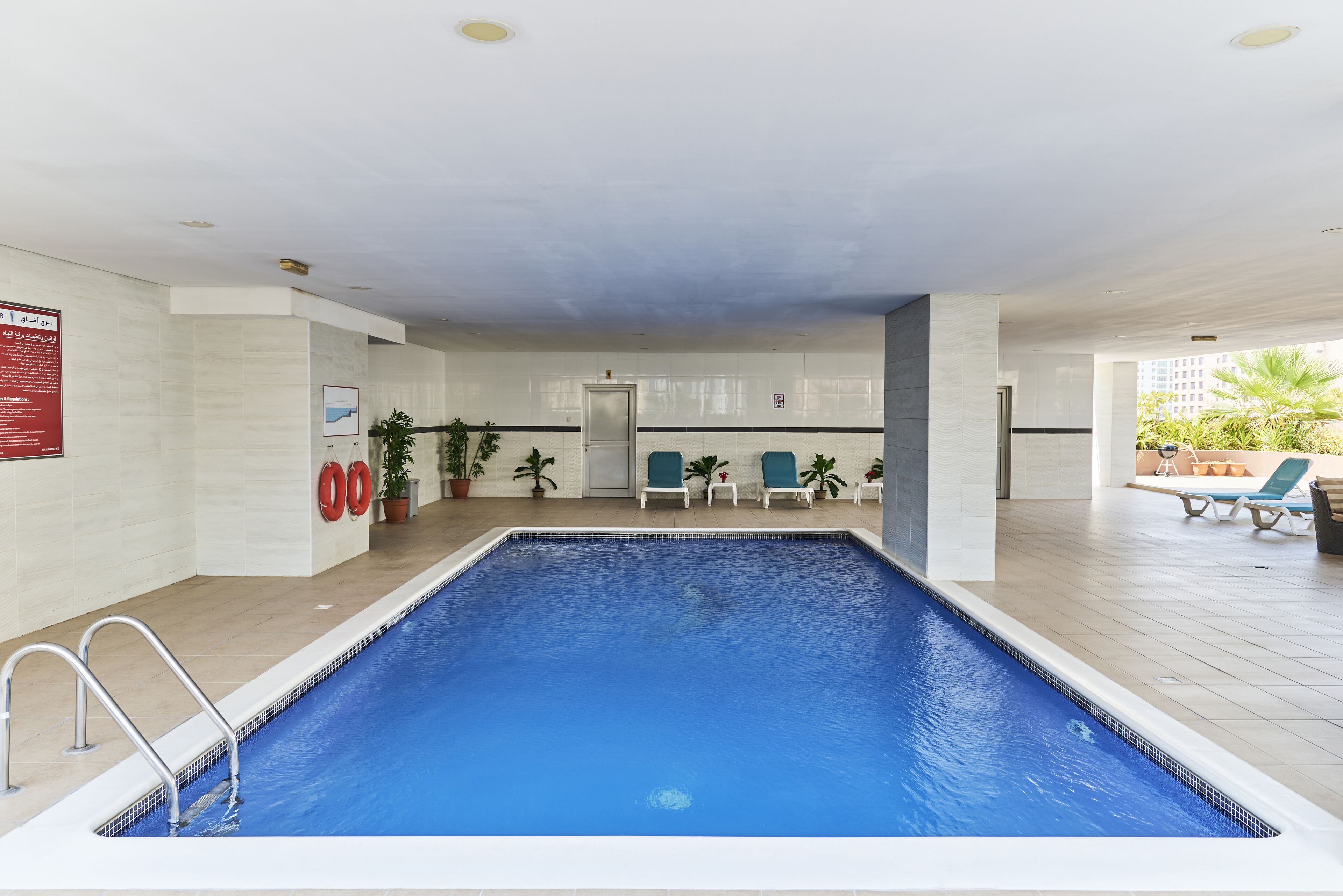 Piscina interna com chão de azulejos, cadeira de salva-vidas e uma porta ao fundo.
