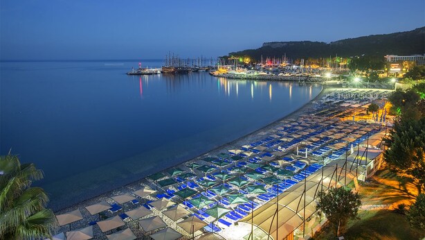 Une plage bordée de rangées de parasols et de chaises longues, une marina avec des bateaux, et une colline parsemée de bâtiments.