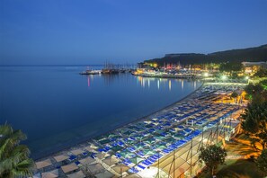 Uma praia com fileiras de guarda-sóis e espreguiçadeiras, uma marina com barcos e uma colina com edifícios.