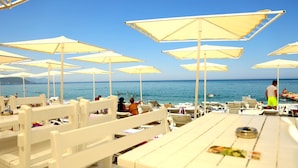 Un restaurant situé en bord de mer, avec des tables et des chaises blanches, des parasols et une vue sur l’océan.