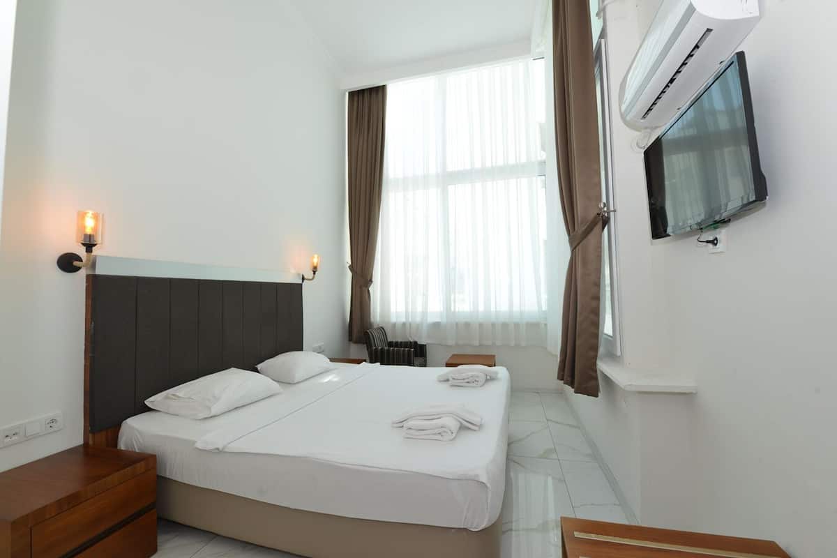 Una habitación de hotel con cama, mesita de noche, lámpara, aire acondicionado y televisor en la pared.
