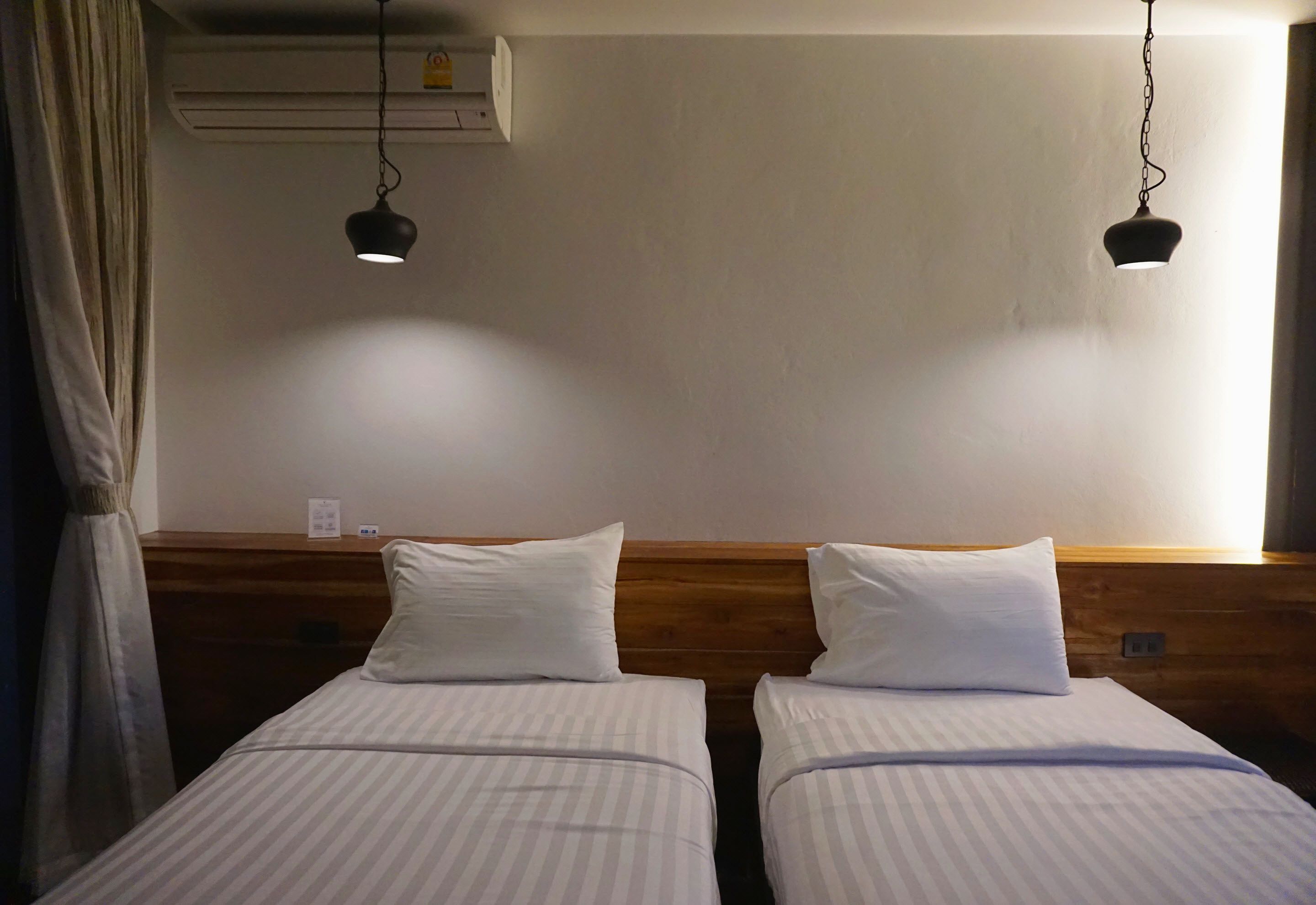 A hotel room with two single beds, wooden headboards, white bedding, and two hanging pendant lights.