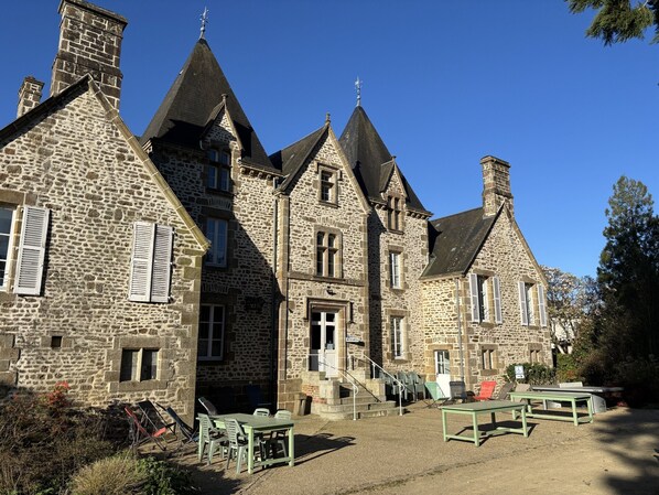 A stone building with multiple chimneys and a staircase leading to the entrance. There are outdoor seating areas with tables and chairs. - Château du Bourg (St Denis de Gastines)