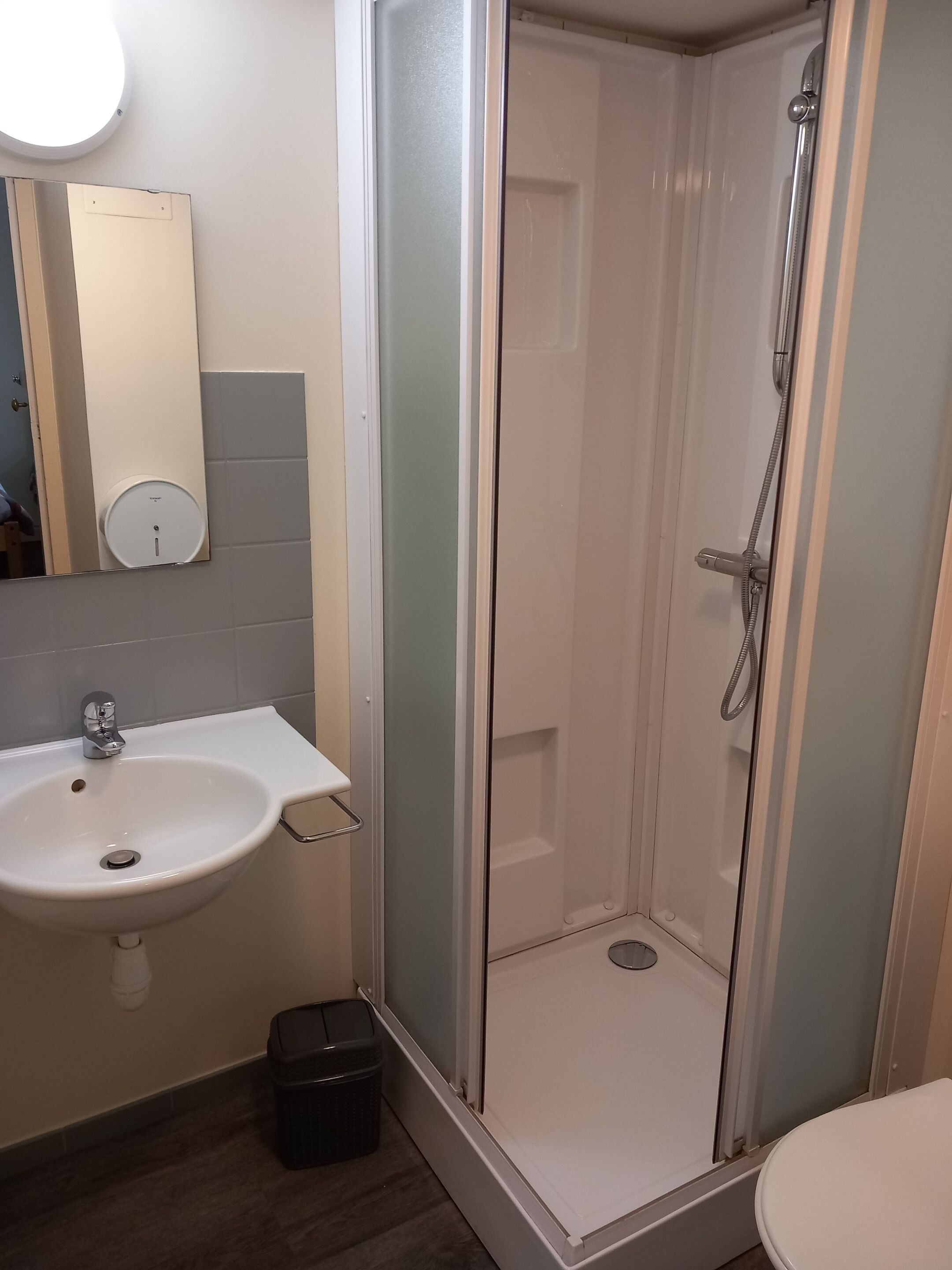 Een badkamer met een wit wastafel, een spiegel, een douche met glazen wanden en een prullenbak.