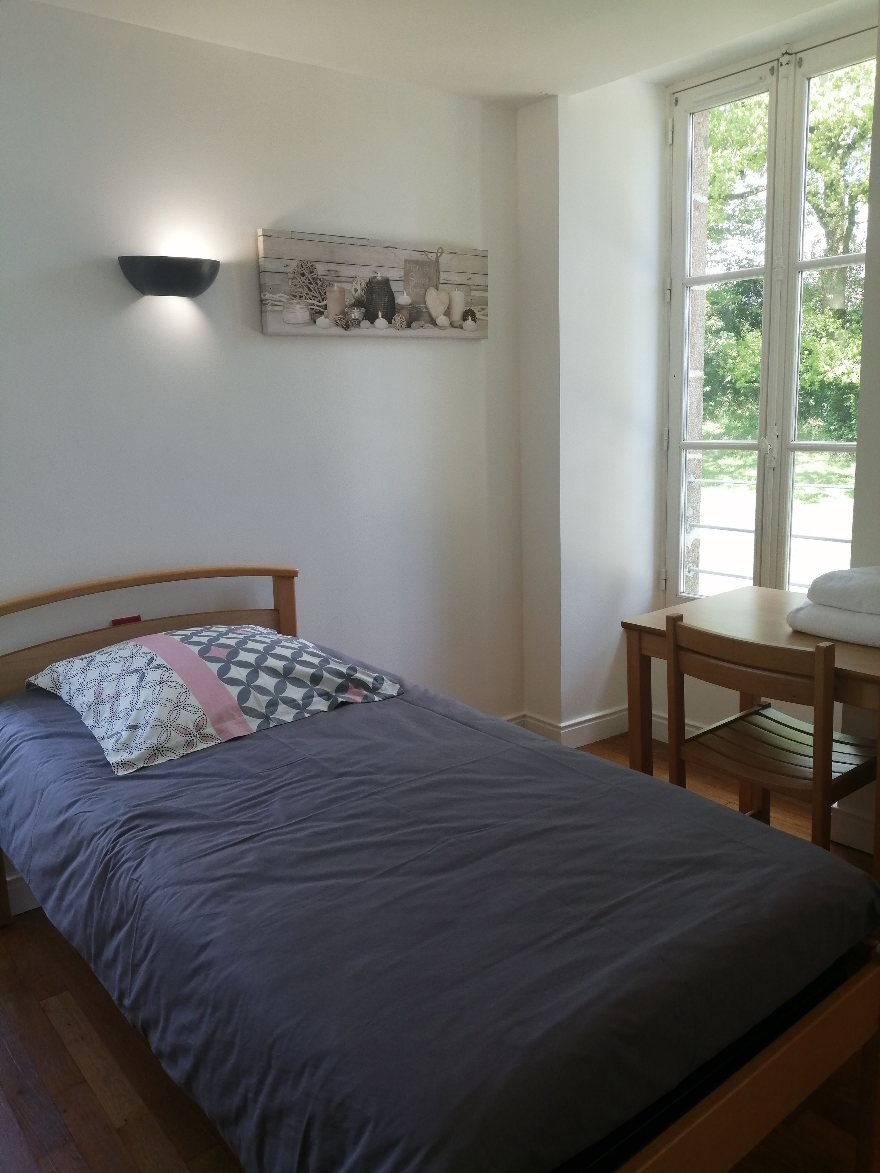 A bedroom with a bed, a wooden headboard, a wall-mounted light, a small table, and a window with a view of greenery.
