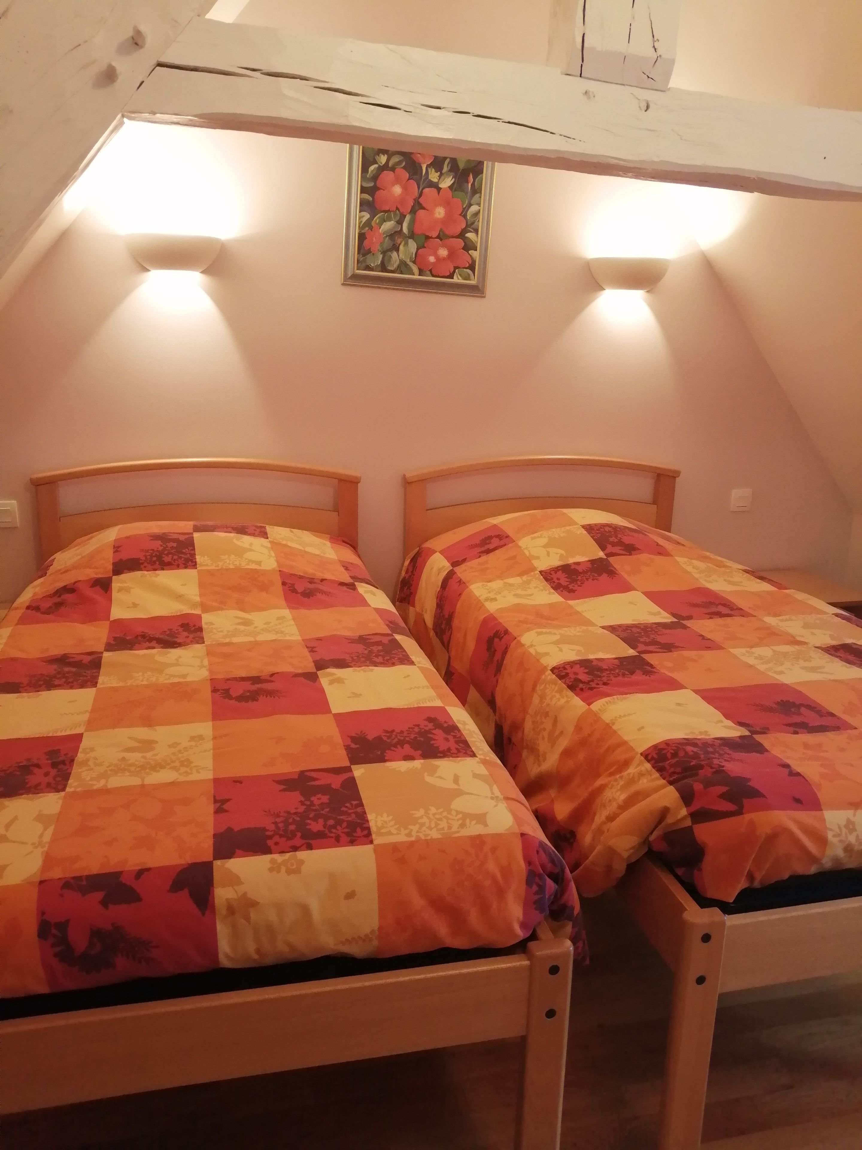 Ein Schlafzimmer mit zwei hölzernen Betten, jedes mit einer karierten Steudecke, unter einer geneigten Decke mit einem Blumenbild.