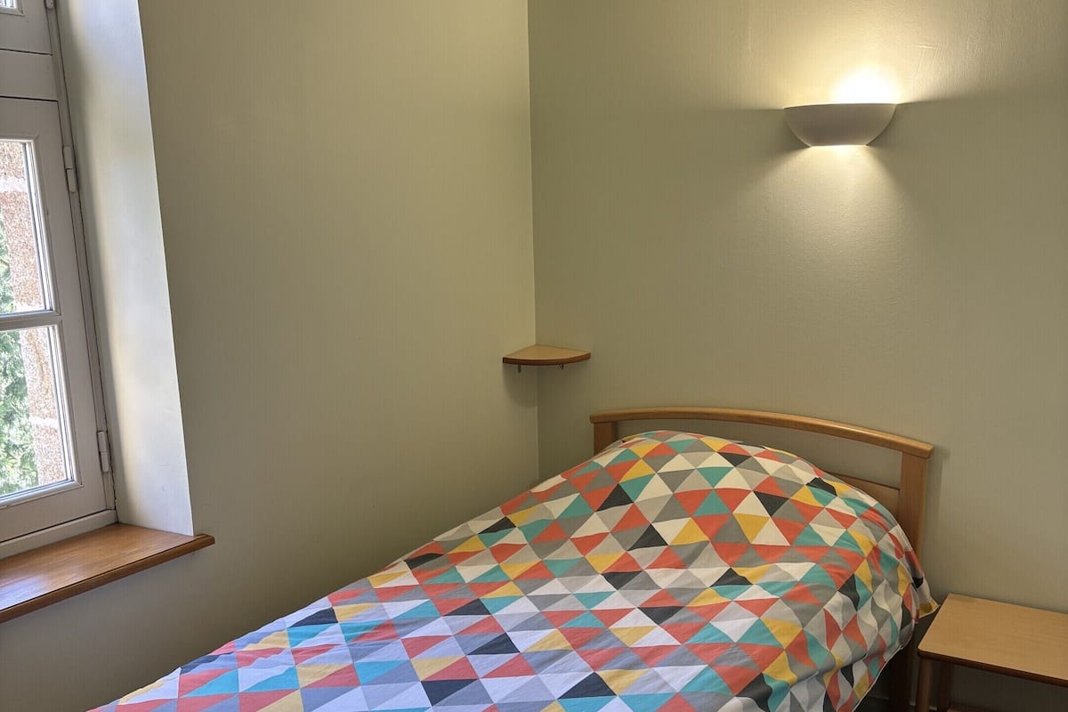 Een net opgemaakt bed met een kleurrijk, geometrisch beddegoed, een houten hoofdbord, een nachtkastje en een raam met uitzicht op groen.