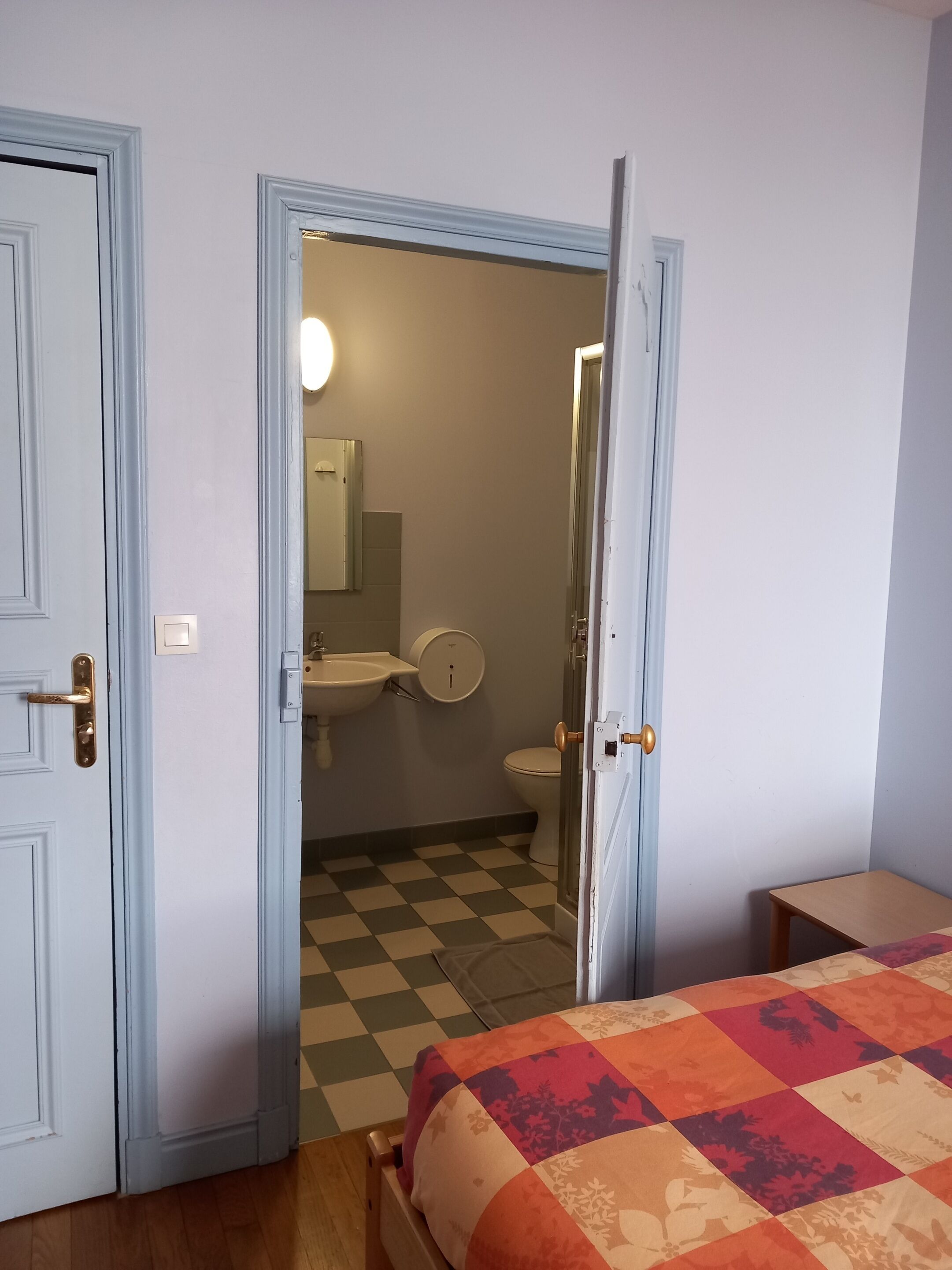 Un dormitorio con suelo a cuadros, una cama con colcha y un baño visible a través de una puerta abierta.