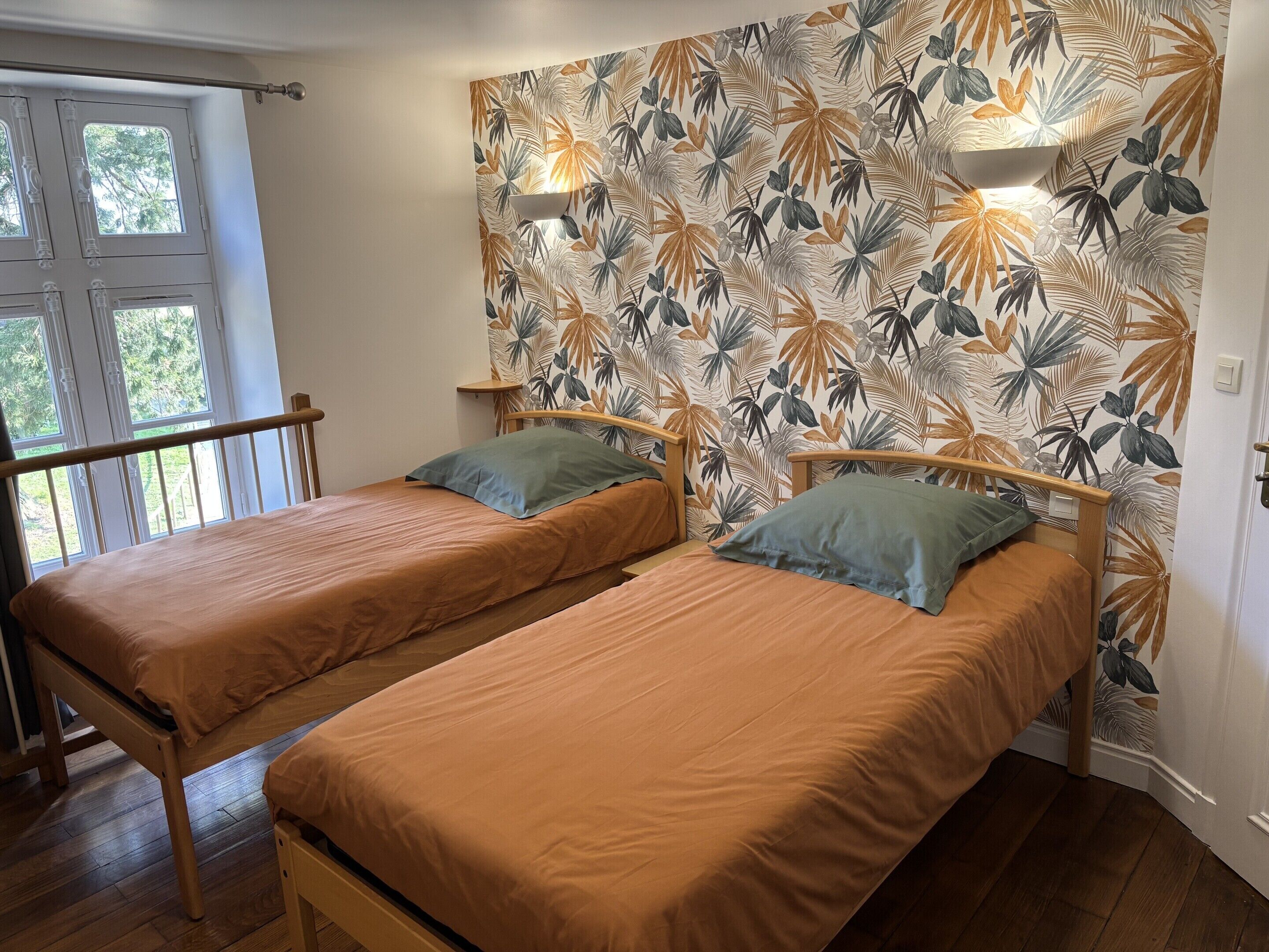 Habitación con dos camas individuales, una ventana con cortinas y un diseño de hojas tropicales en la pared.