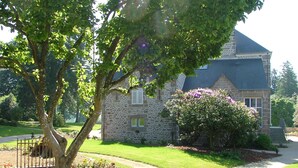 Une maison en pierre avec une pelouse bien entretenue, un arbre au premier plan et un ciel dégagé.