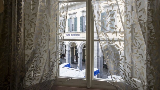 A window with sheer curtains, a view of a building with arches and a sign reading "YEARNO SIGNORELLI". - Appartamento Il Teatro (Cortona)
