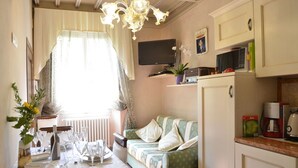 A cozy living room with a dining table set for four, a sofa, a television, and a kitchen area with a coffee maker. - Appartamento Il Teatro (Cortona)