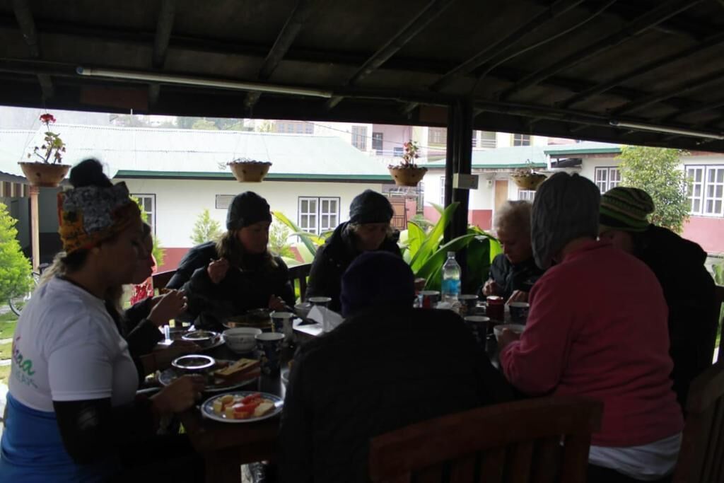 Un grupo de personas cenando en un restaurante al aire libre con mesas y sillas, rodeado de plantas colgantes y un edificio al fondo.
