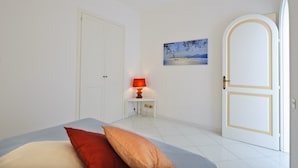 Un dormitorio con cama, almohadas, una mesita de noche con lámpara, un cuadro en la pared y una puerta.