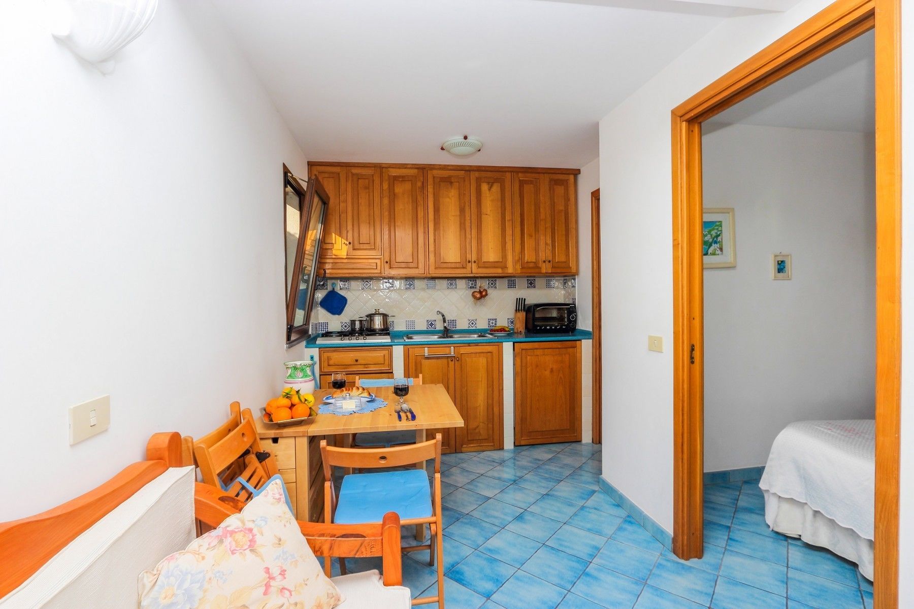A compact kitchen with wooden cabinets, a blue tiled floor, a dining table with chairs, and a bed in the adjacent room.