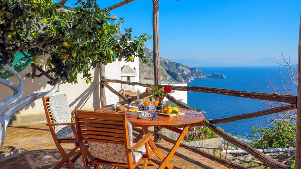 A wooden table with a chair, a small vase with flowers, and a plate of food on a terrace overlooking the sea. - Casa Emotion (Praiano)