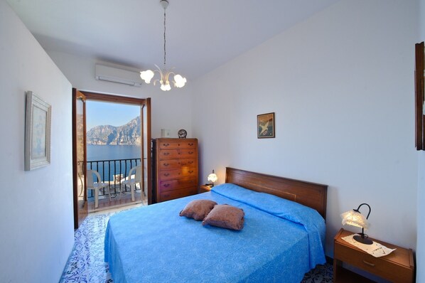 A bedroom with a bed, a dresser, a nightstand, a lamp, and a view of the sea through a balcony door. - Casa Limoncello (Praiano)