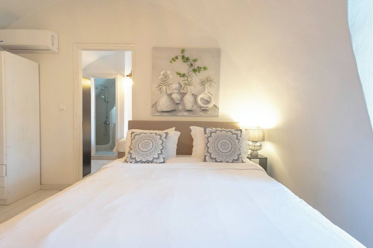 A bedroom with a bed, two bedside lamps, a painting above the bed, and a door leading to a bathroom.