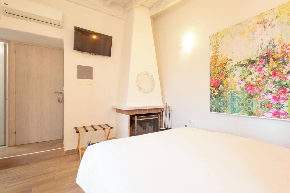 A bedroom with a large bed, a fireplace, a TV mounted on the wall, and a floral painting.
