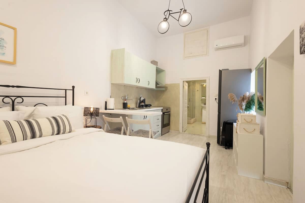 A bedroom with a bed, bedside table, lamp, and a view into the adjacent kitchen.