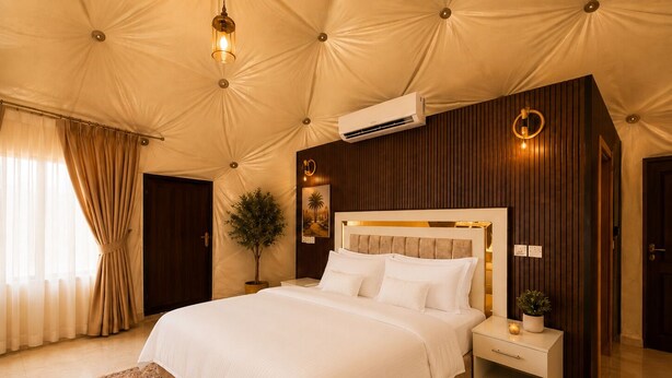 A spacious bedroom with a large bed, a wall-mounted air conditioner, a bedside table with a lamp, and a decorative plant. - Hasan Zawaideh Camp (Wadi Rum)