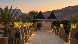 A paved pathway leading to a building with a pyramid-shaped roof, flanked by palm trees and stone walls with illuminated panels. - Hasan Zawaideh Camp (Wadi Rum)