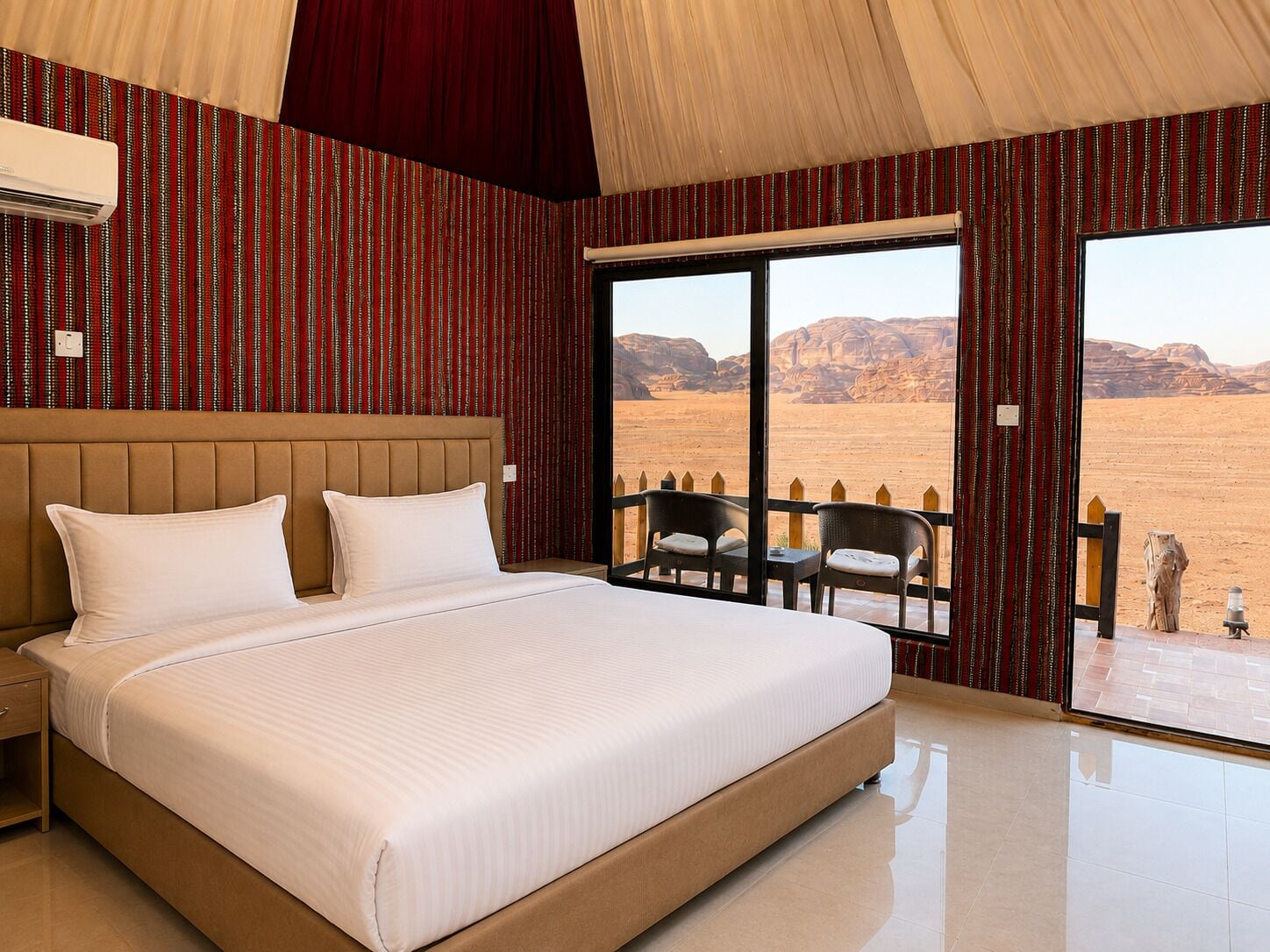 A bedroom with a large bed, a headboard, a window with a view of a desert landscape, and a balcony with chairs.